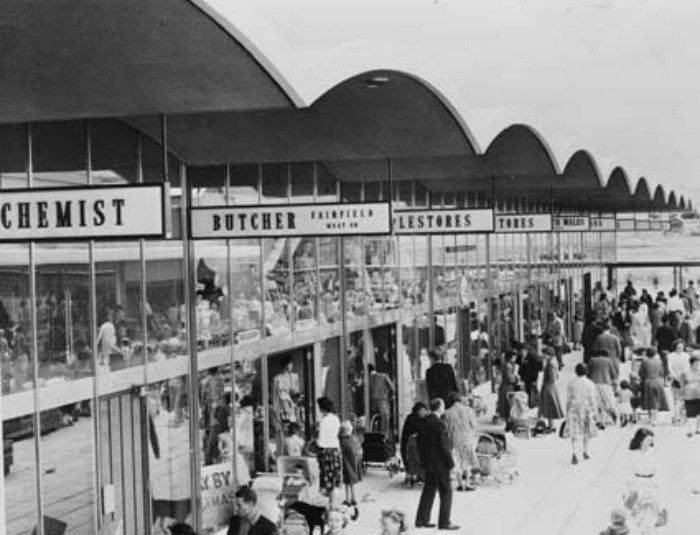 Black and white photo of a busy mid-20th century shopping arcade, featuring storefronts such as a chemist and butcher, with crowds of shoppers walking along the walkway.