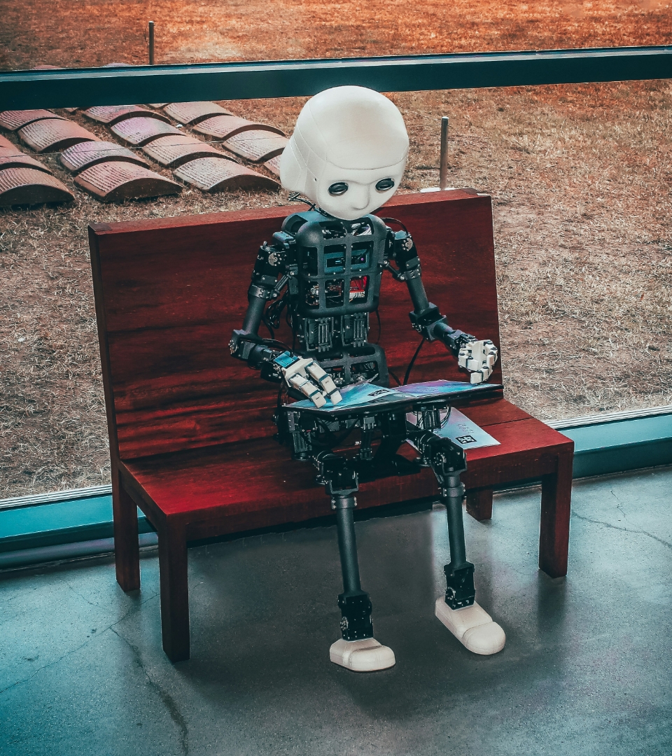 A humanoid robot with a white, childlike face and exposed mechanical components sits on a wooden bench, holding a book in its hands.