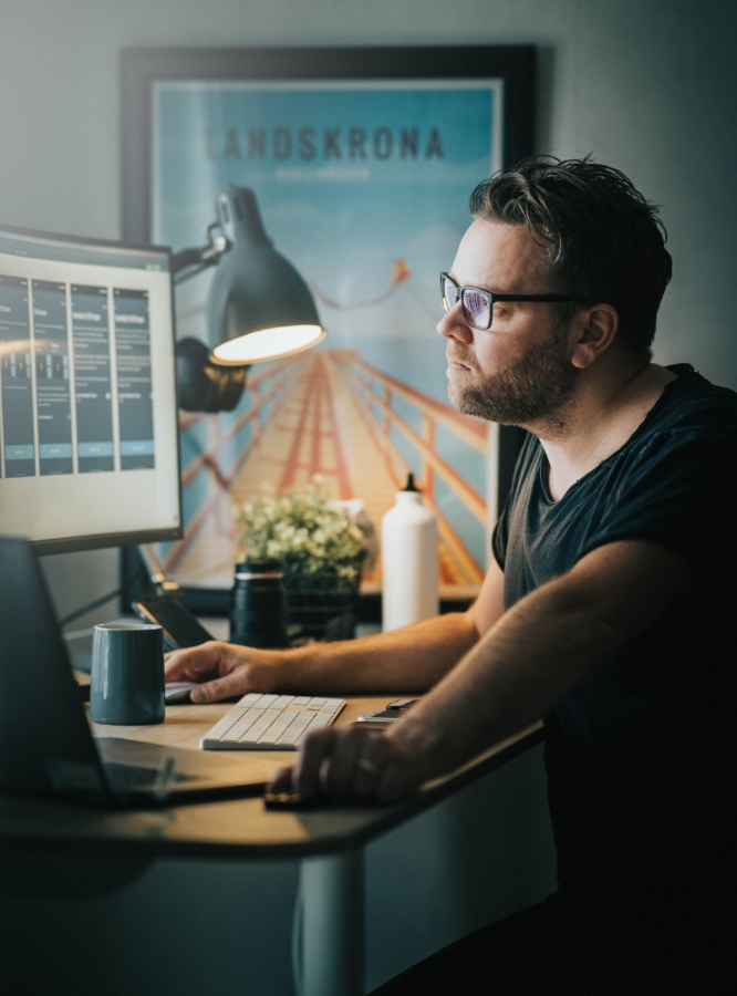 Man wearing glasses focused on a computer screen in a dimly lit workspace, with a "Landskrona" poster in the background.