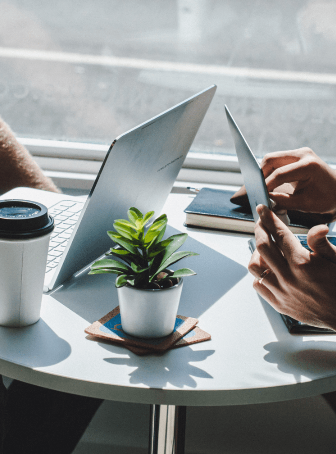 Two professionals working on laptops at a sunlit table with a plant and coffee, symbolising digital collaboration and modern strategy.